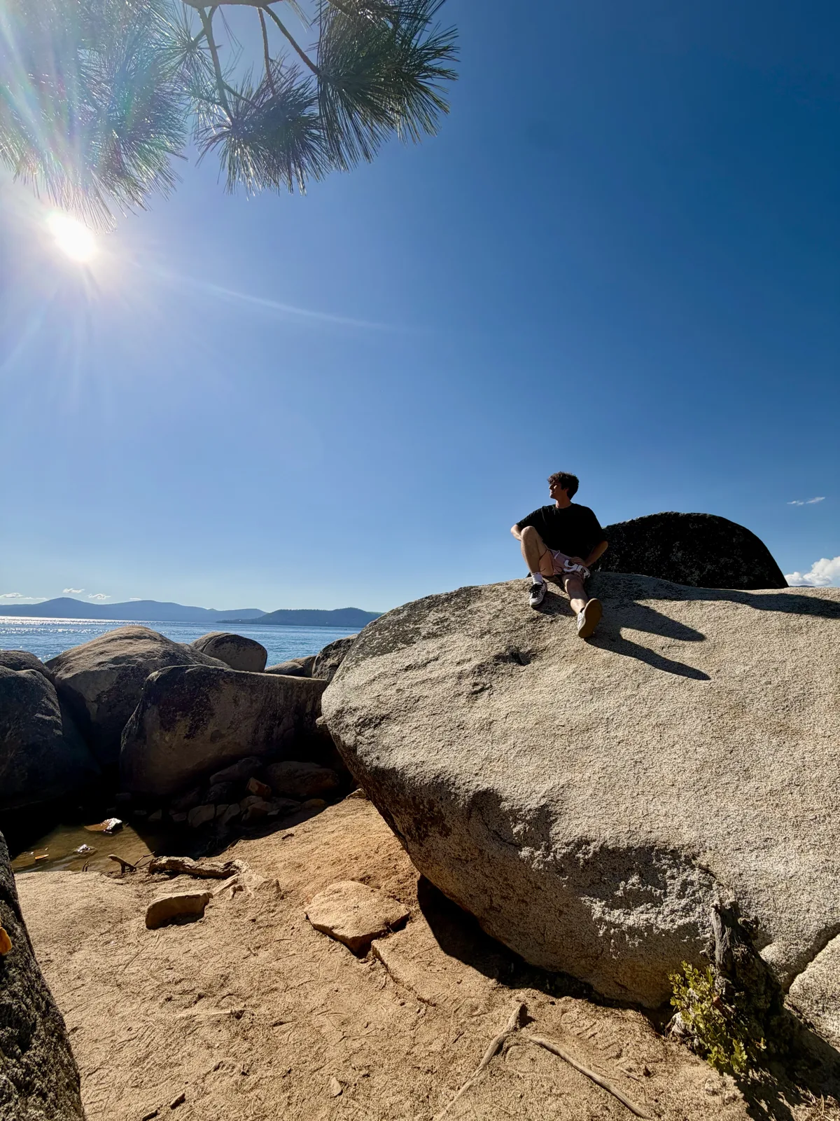 A graduate sitting on a granite boulder above an alpine lake, sun flaring through pine branches.